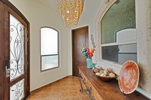 Un corridoio con una porta in legno, un'ampia finestra, un lampadario decorativo e una panca in legno con un vaso di fiori.