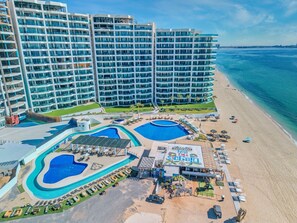 Um hotel com várias piscinas, uma área de praia e um edifício grande com muitas varandas.