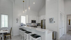 Uma cozinha moderna com armários brancos, uma ilha central com banquetas, eletrodomésticos de aço inoxidável e iluminação pendente.