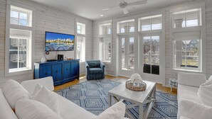 A spacious living room with a large TV, blue and white patterned rugs, and a view of the city through large windows.