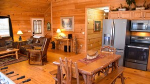 A cozy log cabin interior with a wooden dining table, chairs, a sofa, a fireplace, and a kitchen area with appliances.