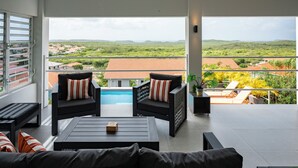 A balcony with a view of a swimming pool and distant hills, furnished with black sofas and striped cushions.