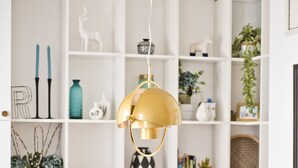 Uma sala de jantar com uma mesa redonda, cadeiras de madeira, uma luminária pendente dourada e um móvel de estantes branco com diversos objetos decorativos.