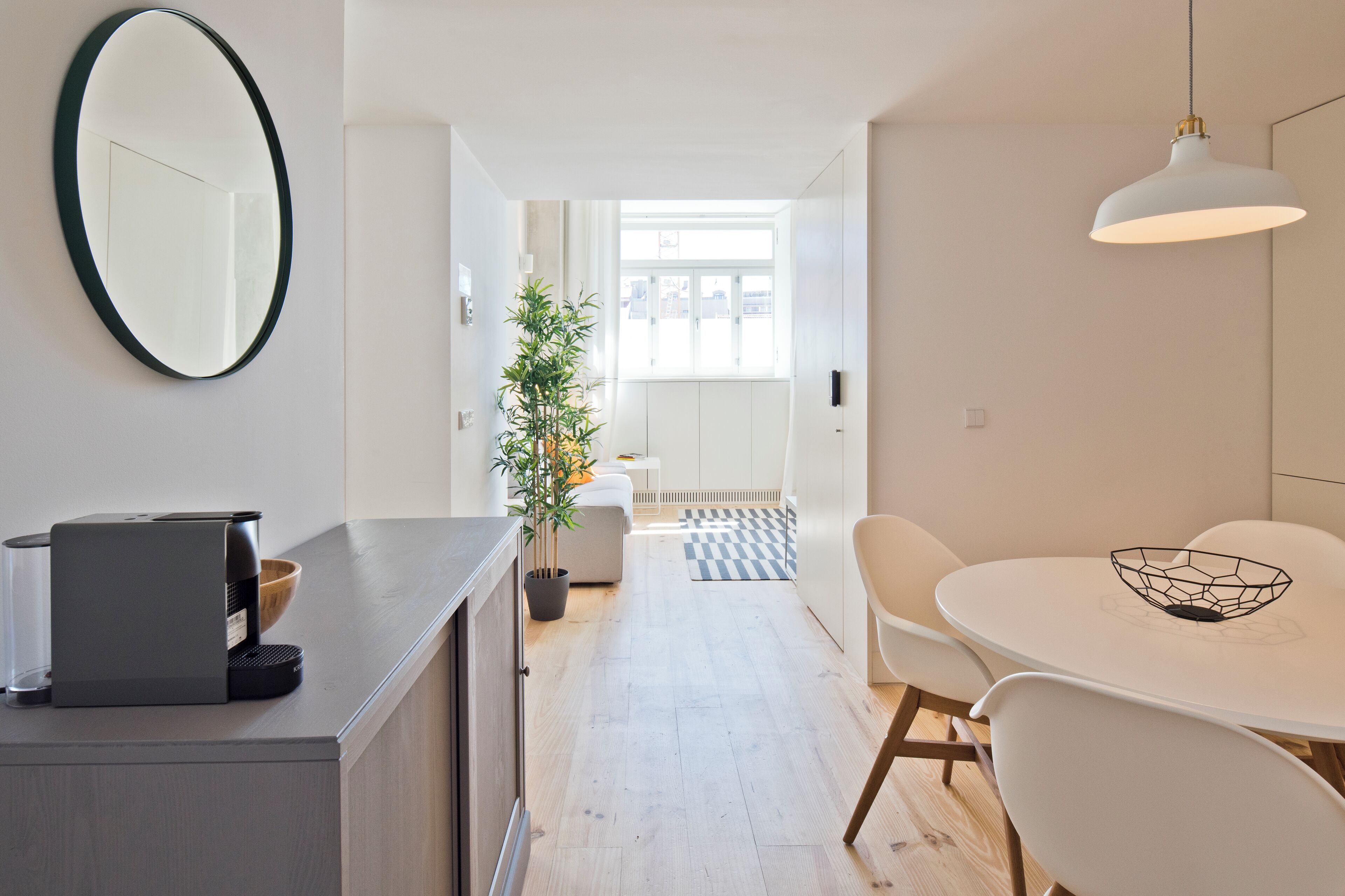 A modern kitchen with a coffee machine, a dining area with a white table and chairs, and a large mirror on the wall.