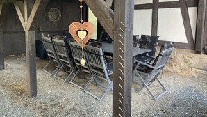 An outdoor seating area with metal chairs and tables under a wooden structure with a sloped roof.