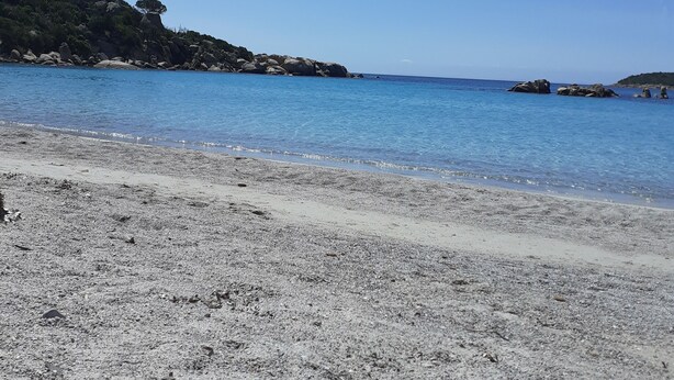 A beach with clear blue water, pebbly shore, and rocky outcrops.
