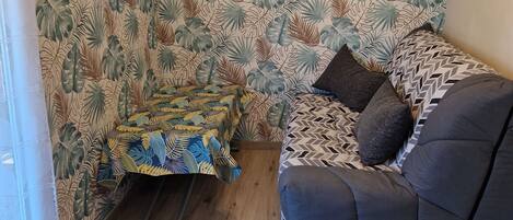 A cozy room with a patterned wall, a window with curtains, a ceiling light fixture, a sofa with patterned cushions, and a small table with a lamp.