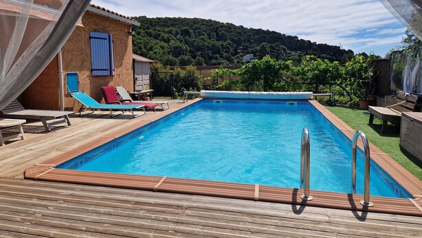 Una piscina rectangular con muelle de madera, rodeada de sillones de descanso y un edificio pequeño con una puerta azul.