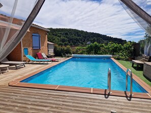 A rectangular swimming pool with a wooden deck, surrounded by lounge chairs and a small building with a blue door.