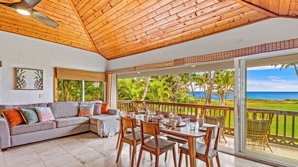 Een ruime woonkamer met houten plafond, een grijze bank met kleurrijke kussens, een eettafel met stoelen en een grote schuifglazen deur die leidt naar een balkon met uitzicht op zee en palmbomen.