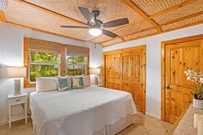 A bedroom with a large bed, wooden doors, a ceiling fan, and a window with a view of greenery.