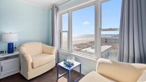 Hotelkamer met een groot raam dat uitzicht biedt op strand en gebouwen, een beige fauteuil, een klein tafeltje met een decoratief object, en een nachtkastje met een lamp en een fles.