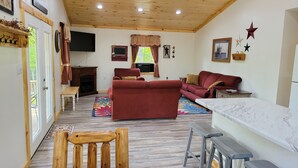 A cozy living room with a fireplace, a red sofa, a wooden dining table, and a TV mounted on the wall. - The Wildflower Cottage at Mountain Paradise (French Creek)
