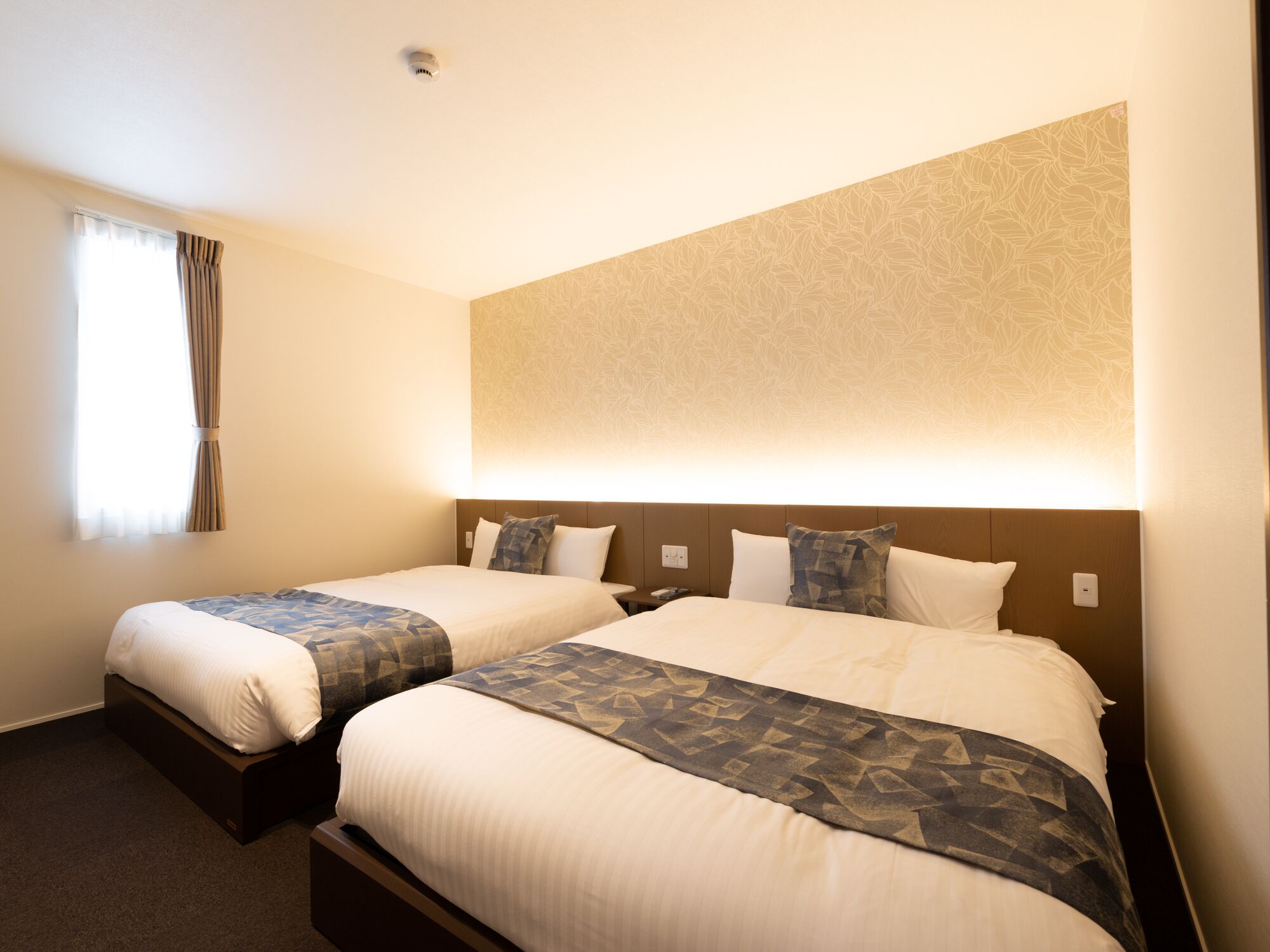 A hotel room with two beds, a large wall-mounted light, and a window with curtains.