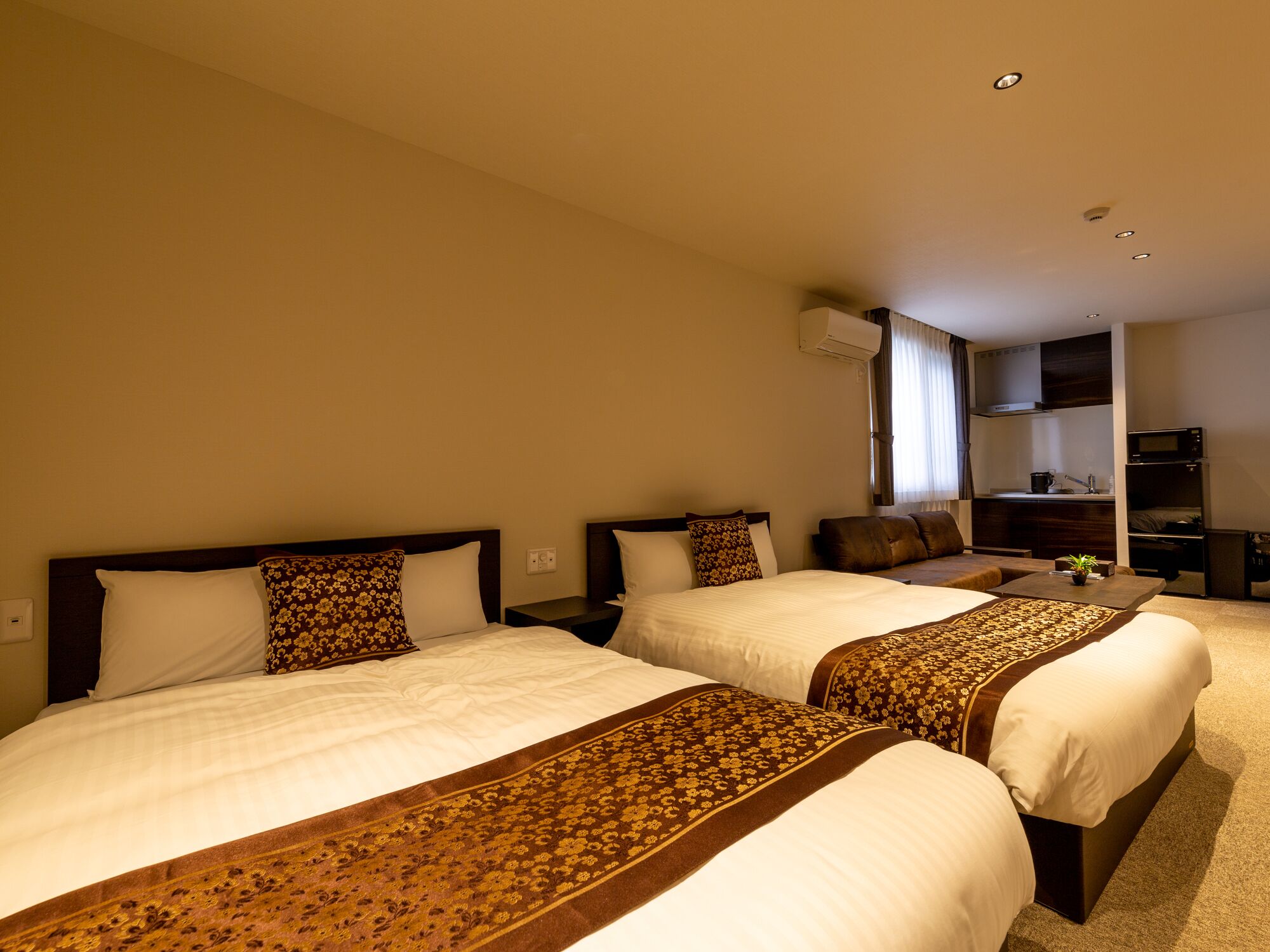 A hotel room with two beds, a sofa, a kitchenette, and an air conditioning unit.