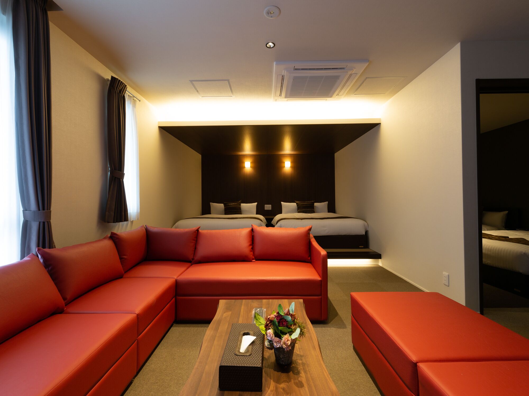 A modern hotel room with a sectional sofa, a wooden coffee table with a vase, and a separate bed area with a headboard.