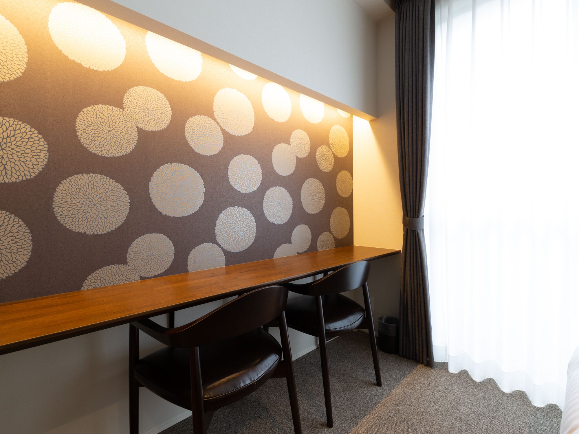 A hotel room with a long wooden desk, two black chairs, and a wall with a circular pattern.