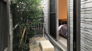 Un espacio exterior reducido con valla de madera, un pequeño jardín con piedras y una puerta que da a una habitación con cama y ventana.