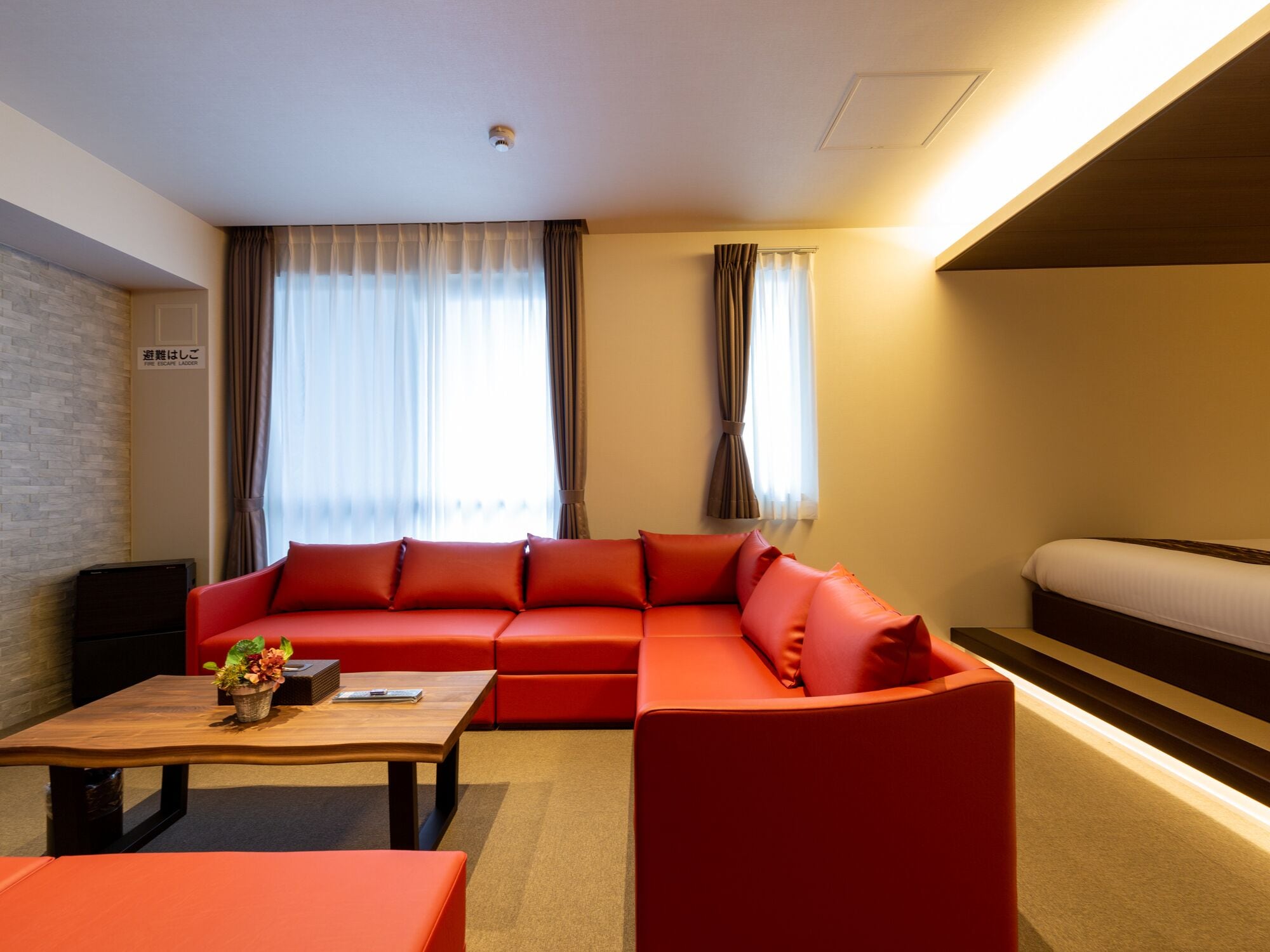A modern hotel room with a sectional sofa, a single bed, a wooden coffee table, and a small side table.