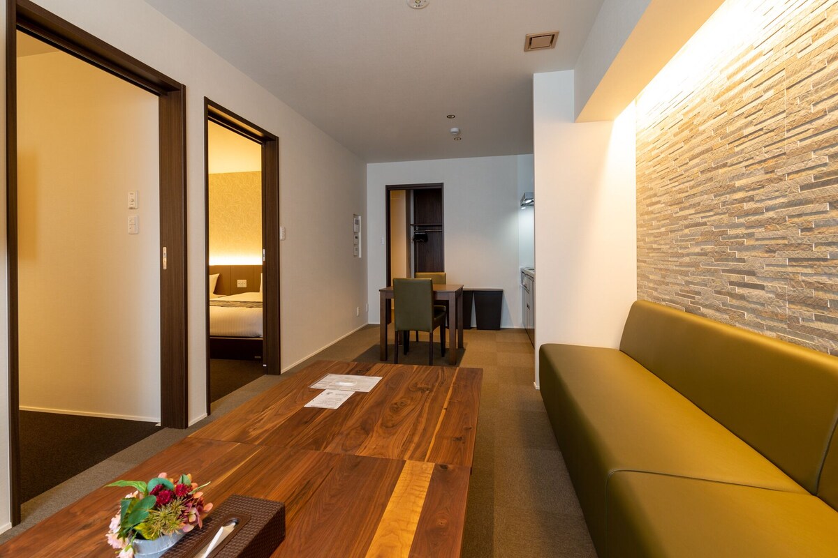 A hotel room with a wooden floor, a green sofa, a wooden coffee table with a vase of flowers, a desk with a chair, and a stone-textured wall.
