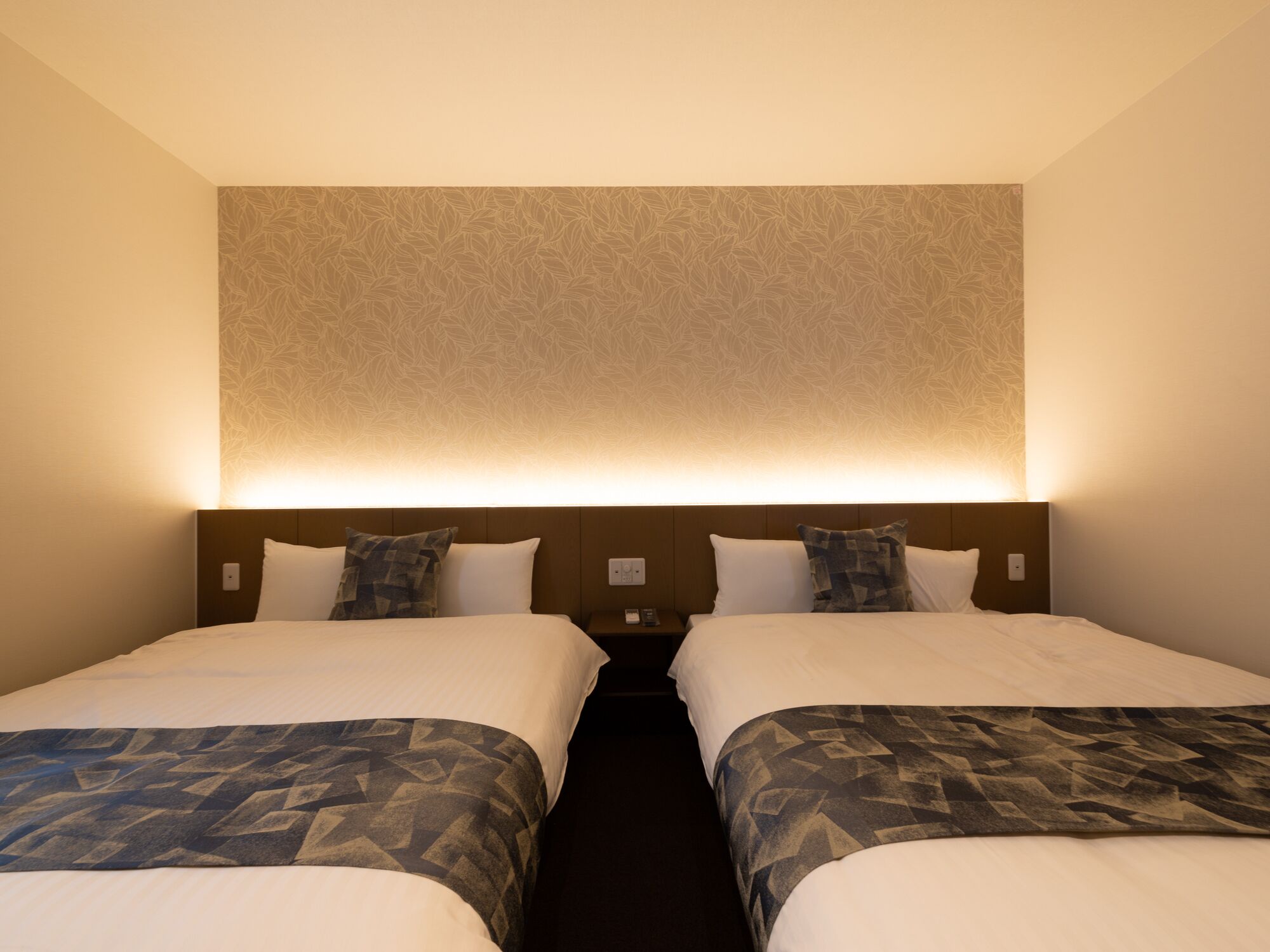 A hotel room with two beds, each with a patterned bedspread and pillows, and a headboard with a textured design.