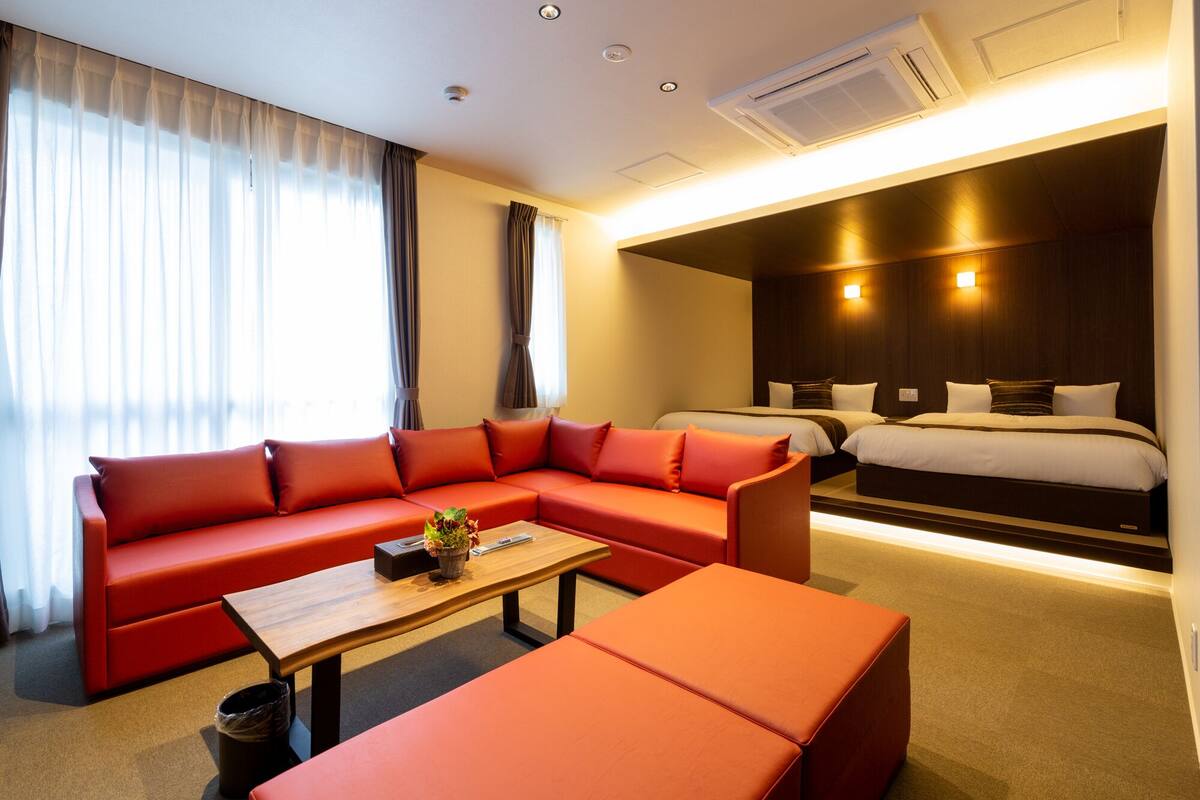 Moderni hotellihuone, jossa on punainen sohva, puinen sohvapöytä, pieni roskakori ja tummanvärinen päätyosalla varustettu sänky.