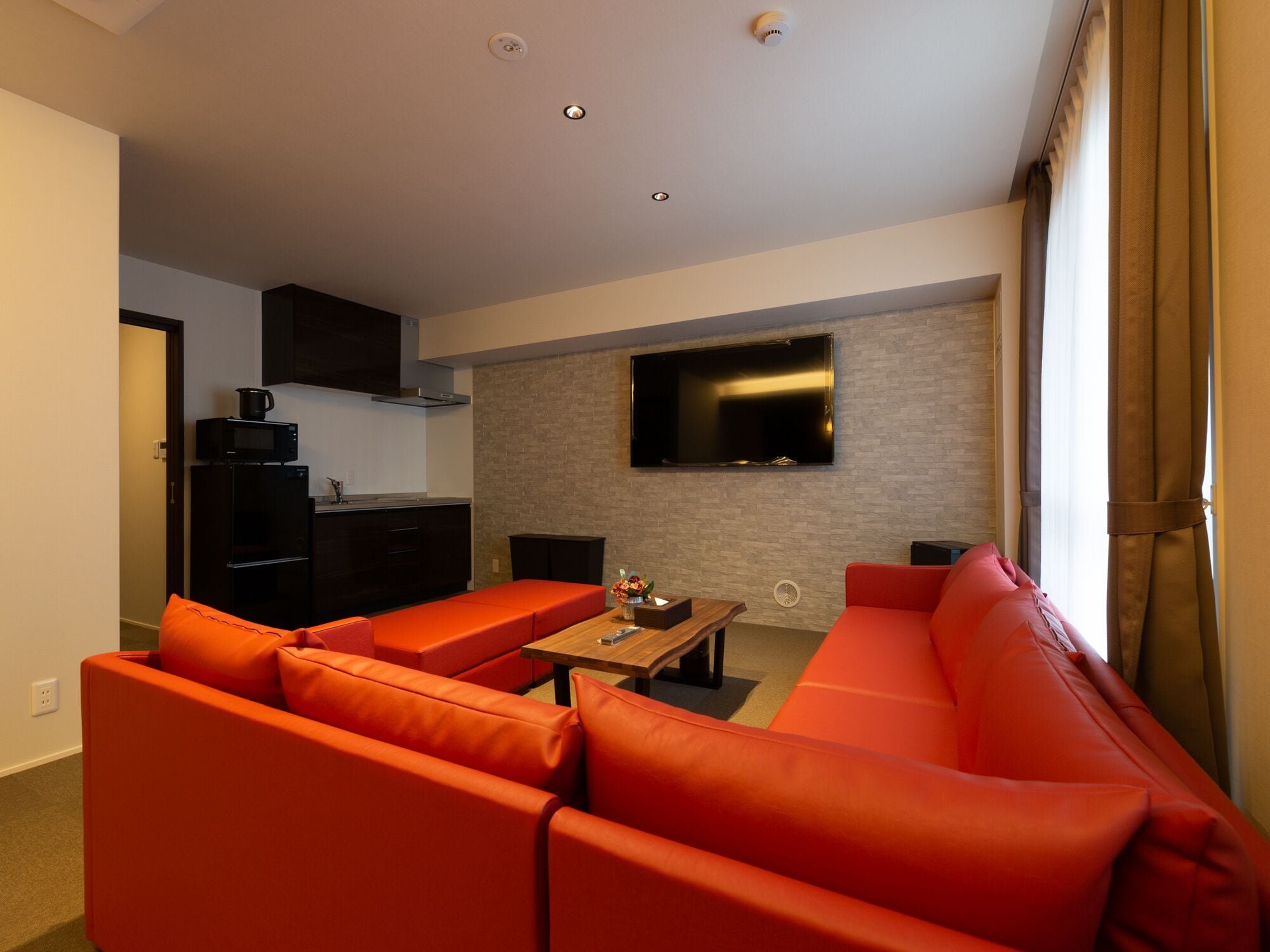 A modern living room with a red sofa set, a flat-screen TV mounted on the wall, and a kitchen area with dark cabinetry and a microwave.