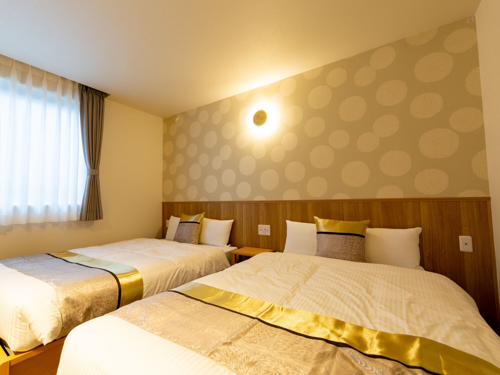 A hotel room with two beds, a wooden headboard, a wall with a polka dot pattern, and a window with curtains.