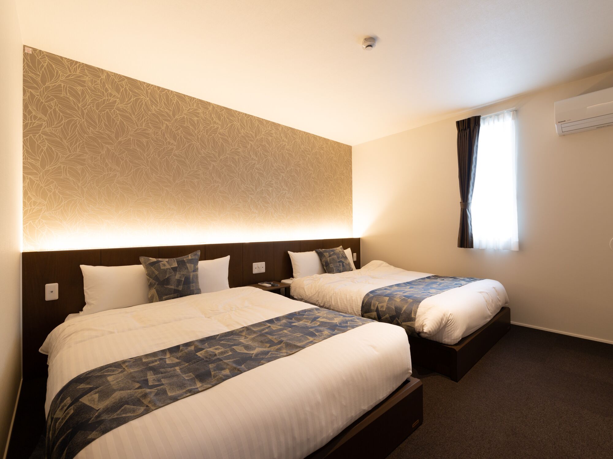 A hotel room with two beds, a patterned wall, a window with curtains, and an air conditioning unit.