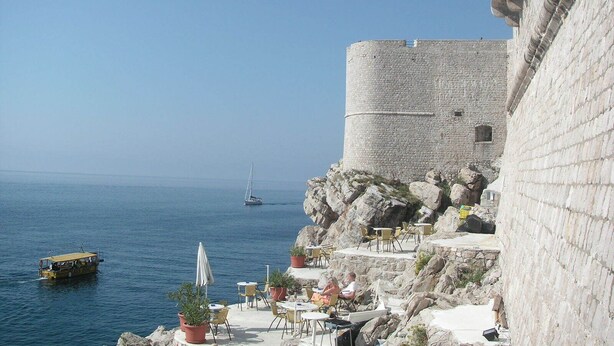 A coastal scene with a stone fortification overlooking the sea, outdoor seating with tables and chairs, and a boat in the water. - Apartment close to the beach and main street (Dubrovnik)