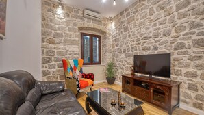 A living room with a stone wall, a leather sofa, a colorful armchair, a wooden coffee table, and a TV on a wooden stand. - Apartment close to the beach and main street (Dubrovnik)