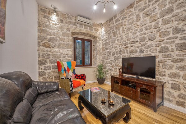Una sala de estar con pared de piedra, un sofá de cuero, un sillón colorido, una mesa de centro de madera y un televisor sobre un mueble de madera.