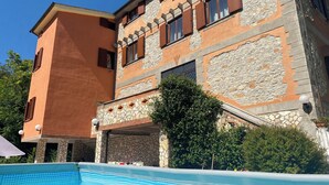 Un edificio de dos pisos con fachada de piedra y contraventanas de madera, una piscina al frente y un cielo azul despejado.