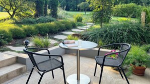A garden patio with two chairs and a table, surrounded by lush greenery and a stone pathway.