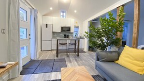 A modern kitchen with white cabinets, a dining area with a table and chairs, a grey sofa with a yellow pillow, and a large potted plant.