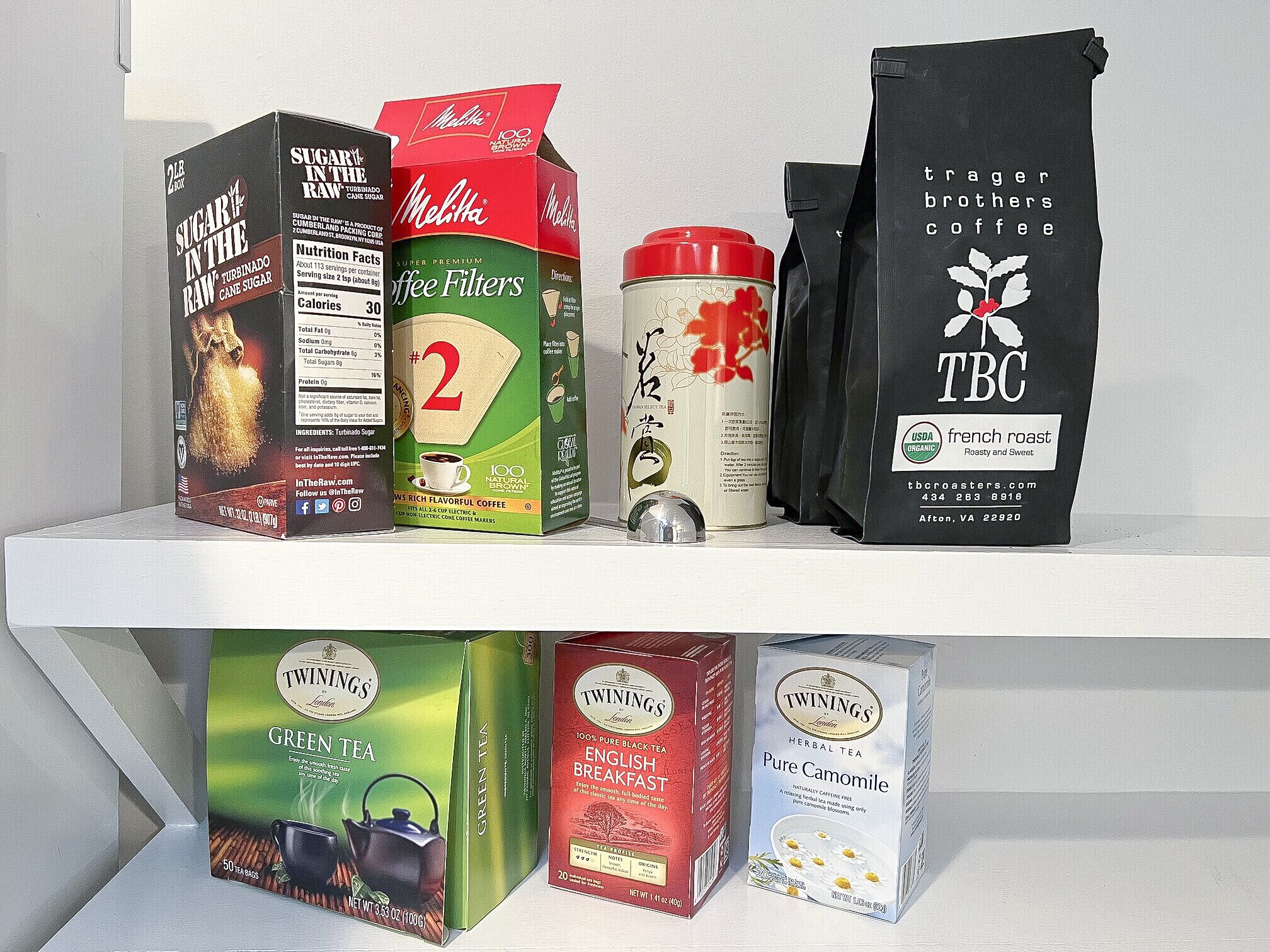 Une étagère contenant divers produits liés au café, notamment une boîte de sucre de canne brut, une boîte de filtres à café Melitta, un récipient de café en grains, un paquet de café torréfié à la française de la marque TBC et des boîtes de thé Twinings.