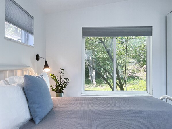 A bedroom with a large window, a bedside lamp, a bed with white and blue bedding, and a potted plant.