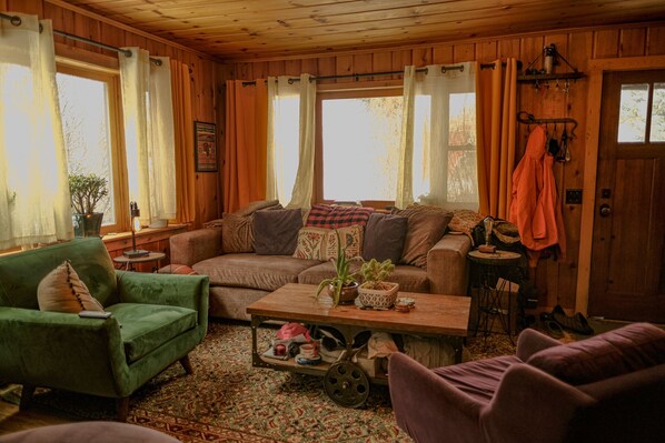 A cozy cabin interior with a green armchair, a sofa, a coffee table with plants, and a rustic wooden floor.