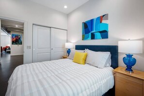 A hotel room with a bed, two bedside tables, a nightstand, a painting on the wall, and a door leading to another room.
