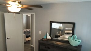 Een slaapkamer met een bed, een dressoir, een spiegel, een plafondventilator en een deur die naar een andere kamer leidt.