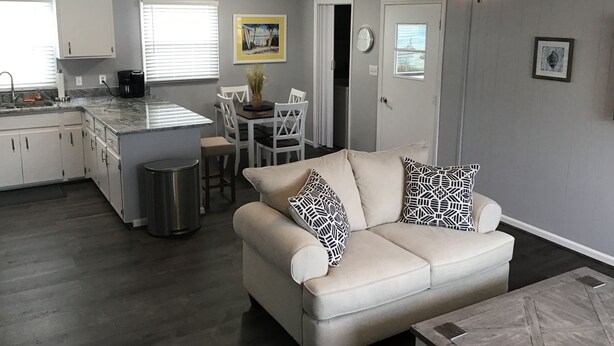 Een moderne keuken met witte kasten, een eethoek met tafel en stoelen, een beige bank met gestreepte kussens, en een plafondventilator.