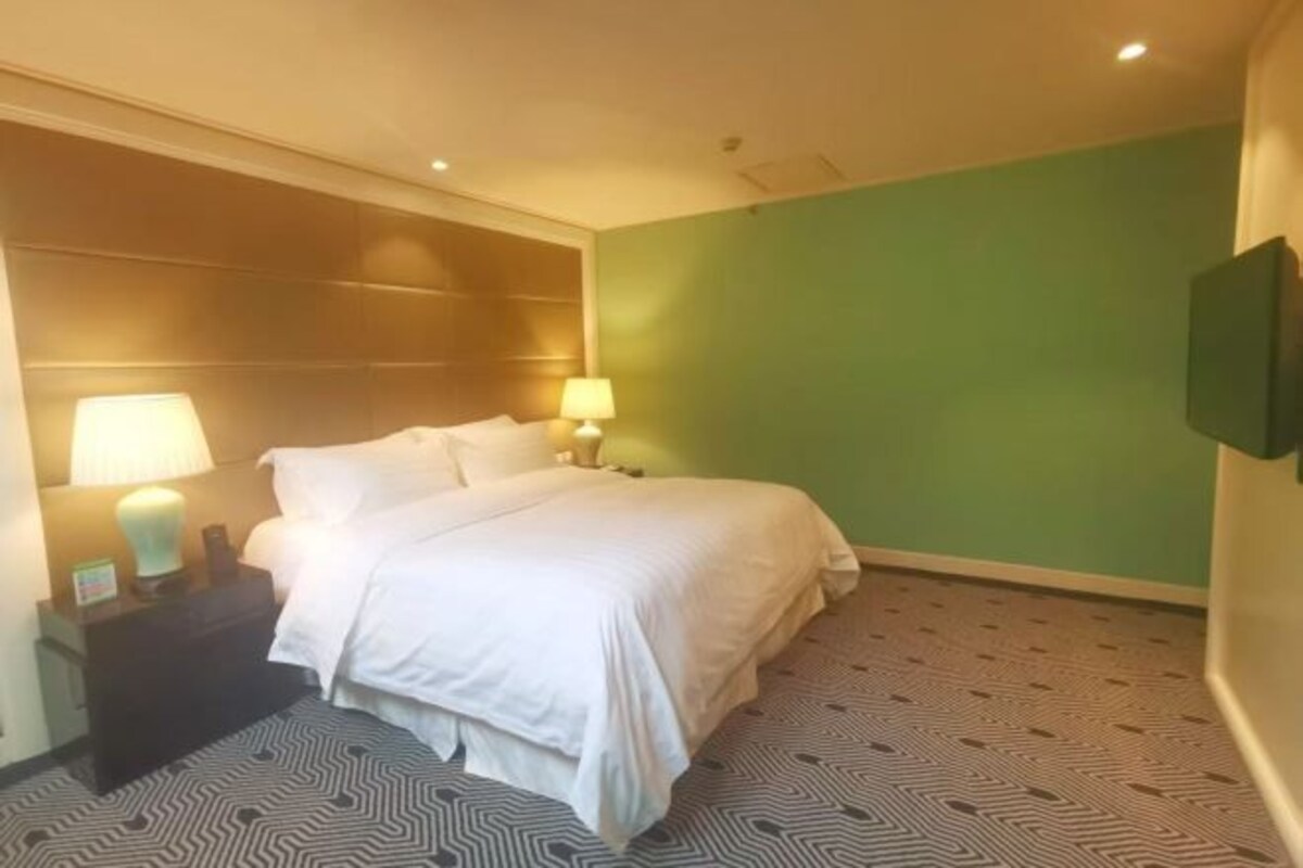 A hotel room with a bed, two bedside lamps, a nightstand, and a green wall.
