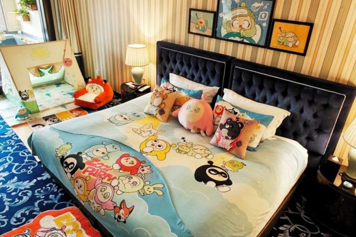 Habitación de hotel con una cama que tiene ropa de cama con personajes de dibujos animados, una mesita de noche con una lámpara y una pared con cuadros.