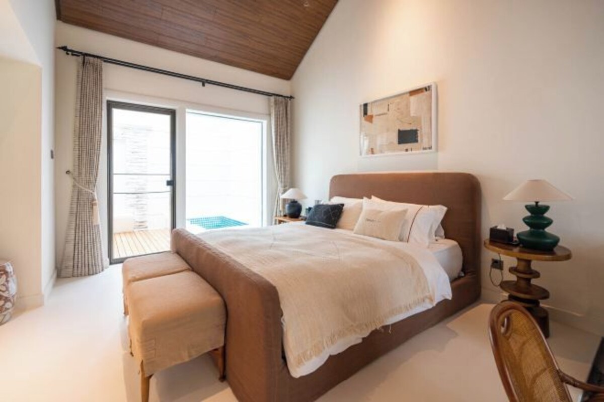 Un dormitorio con una cama grande, un banco, una mesita de noche con lámpara y vistas a un espacio exterior a través de una puerta corredera.