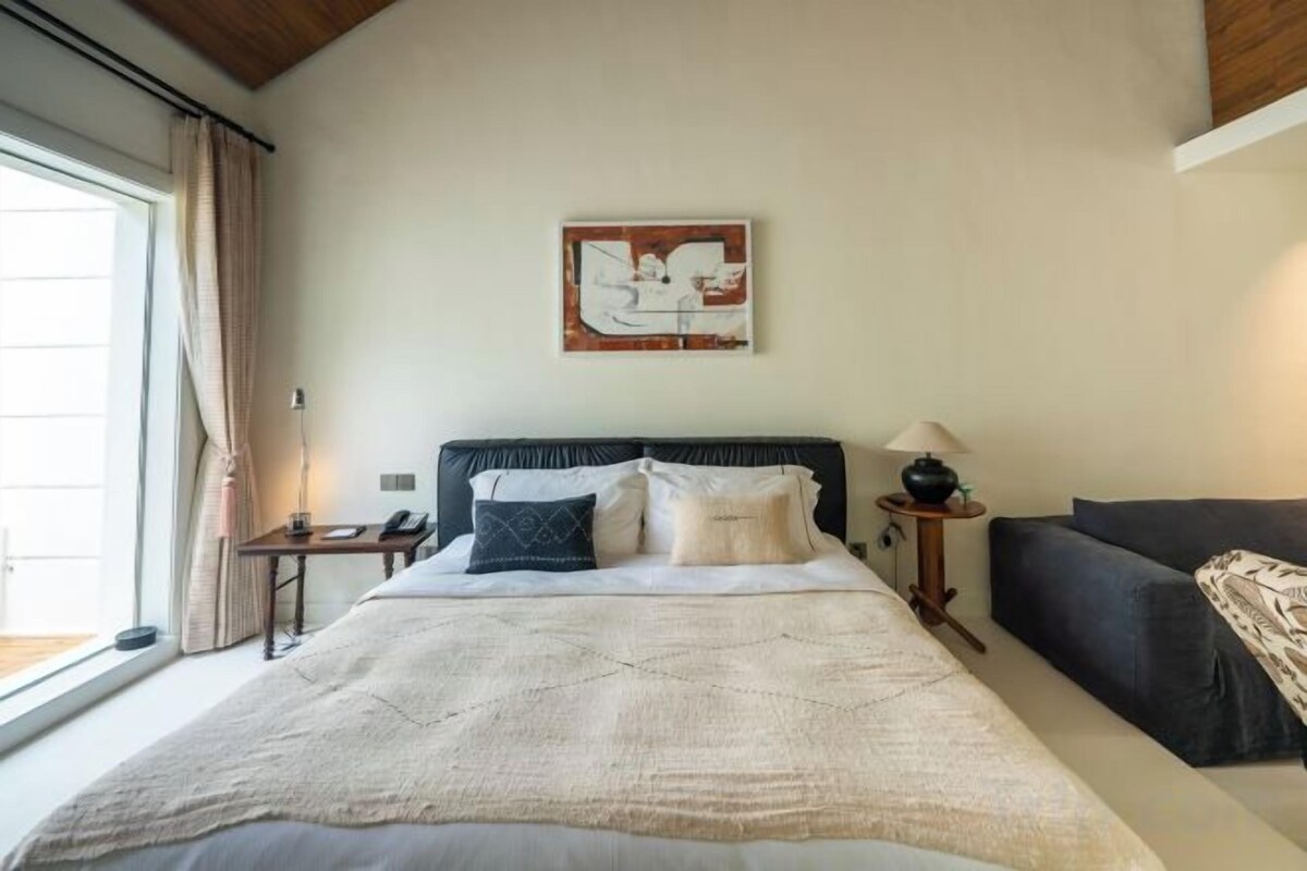 A bedroom with a large bed, a bedside table, a lamp, a sofa, and a window with curtains.