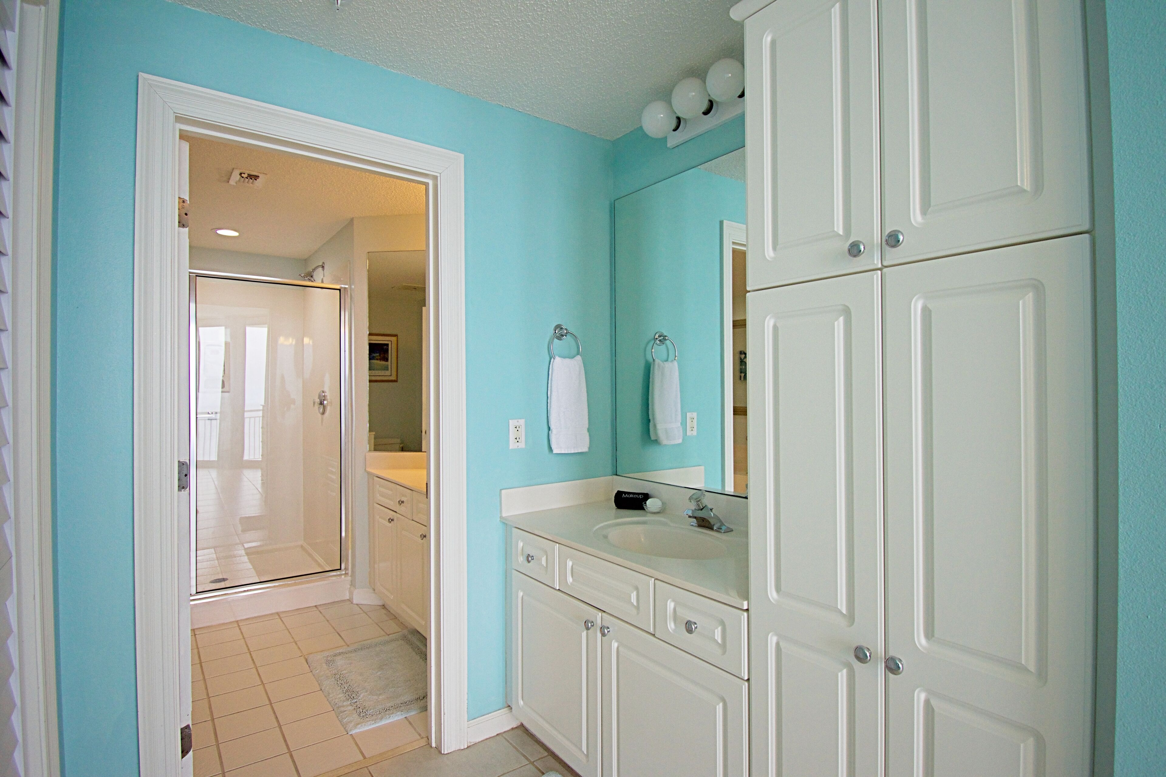Un baño con tocador blanco, un lavabo y un espejo. Hay una puerta que da a otra habitación.
