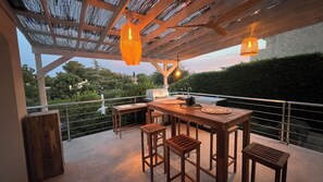 Een terras buiten met een houten tafel en stoelen, een barbecue en hangende lichten.