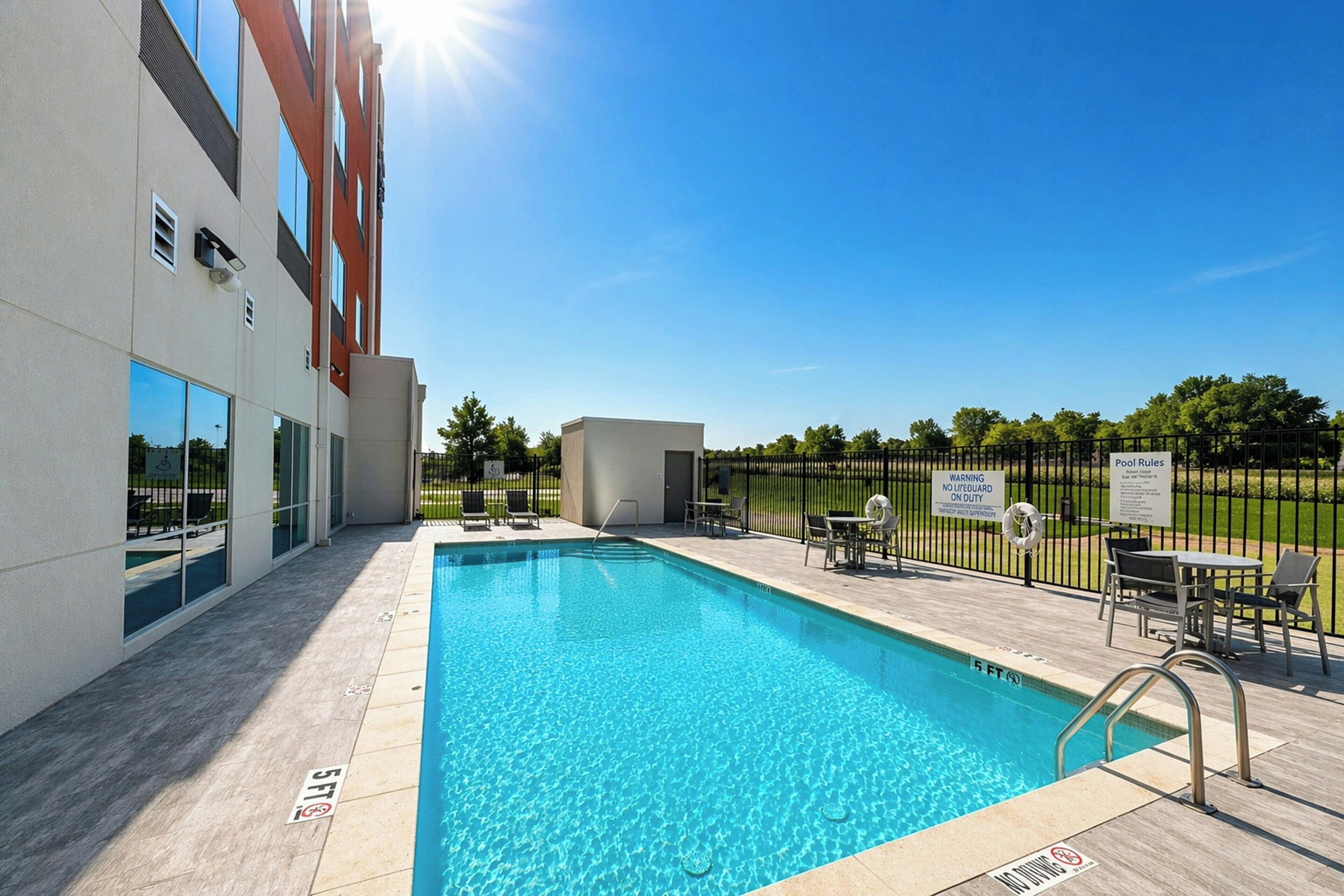 Une piscine à l’eau d’un bleu cristallin, entourée d’une terrasse avec des chaises et des tables, et un bâtiment aux grandes fenêtres en arrière-plan.
