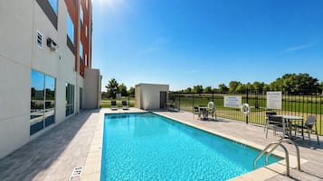 Une piscine à l’eau d’un bleu cristallin, entourée d’une terrasse avec des chaises et des tables, et un bâtiment aux grandes fenêtres en arrière-plan.