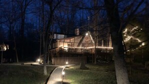 A wooden house with a sloped roof, surrounded by trees. There is a pathway leading to the house, and string lights are visible around the property.